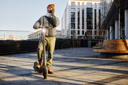 Téléchargez les photos : Jeune homme en vêtements chauds parlant sur téléphone portable tout en conduisant en scooter dans la ville - en image libre de droit