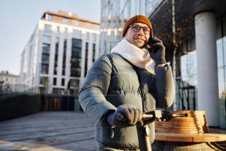 Téléchargez les photos : Jeune homme ayant une conversation sur un téléphone portable tout en conduisant en scooter le long de la rue le jour de l'automne - en image libre de droit