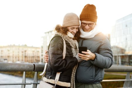 Foto de Joven pareja feliz viendo fotos en el teléfono móvil de pie al aire libre - Imagen libre de derechos
