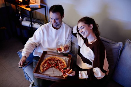Téléchargez les photos : Jeune couple heureux regarder la télévision et manger de la pizza tout en se reposant sur le canapé dans la chambre - en image libre de droit