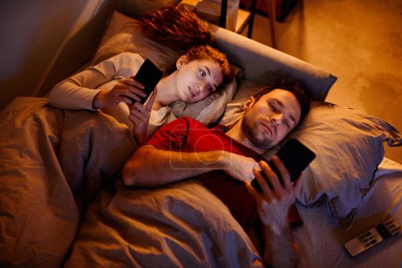 Téléchargez les photos : Vue grand angle du jeune couple à l'aide de gadgets avant de dormir tout en étant couché sur le lit dans la chambre - en image libre de droit