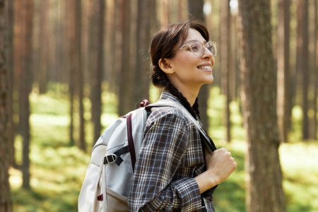 Téléchargez les photos : Jeune femme en lunettes avec sac à dos dans le dos profitant de la nature marchant en forêt - en image libre de droit