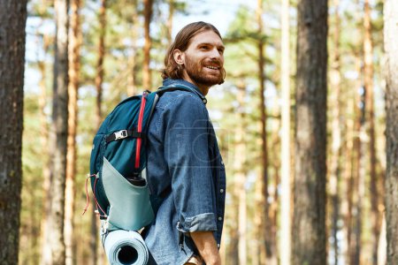 Téléchargez les photos : Jeune homme souriant avec sac à dos randonnée dans la forêt en journée d'été - en image libre de droit