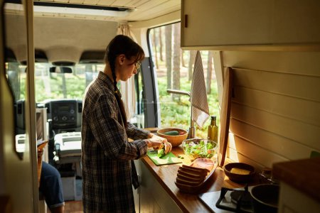 Téléchargez les photos : Jeune femme coupant salade de légumes à la table de cuisine en camping-car au camping dans la forêt - en image libre de droit