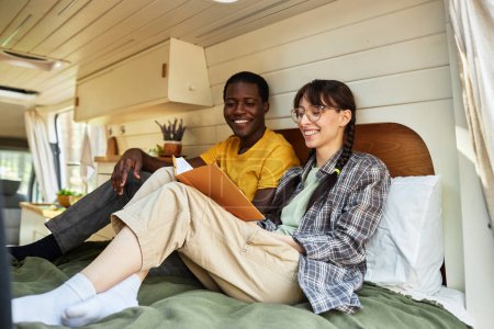 Téléchargez les photos : Heureux couple multiethnique assis sur le lit dans la maison des roues et le livre de lecture ensemble - en image libre de droit