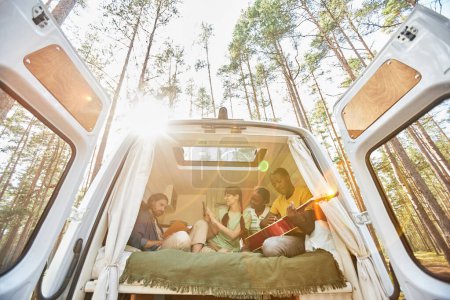 Téléchargez les photos : Groupe d'amis jouant de la guitare assis en camping-car pendant le camping dans la forêt - en image libre de droit