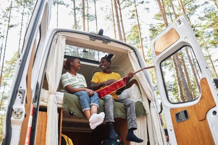Téléchargez les photos : Jeune couple africain jouant de la guitare et chantant des chansons assis en camping-car dans la forêt - en image libre de droit
