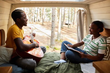 Téléchargez les photos : Jeune couple africain reposant en van jouant de la guitare et lisant un livre pendant le camping dans la forêt - en image libre de droit