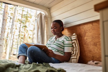 Téléchargez les photos : Jeune fille africaine lisant un livre tout en se reposant sur le lit dans la maison de roues dans la nature - en image libre de droit