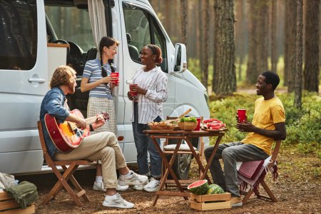 Photo for Young people talking to each other and playing guitar while resting in nature during picnic - Royalty Free Image