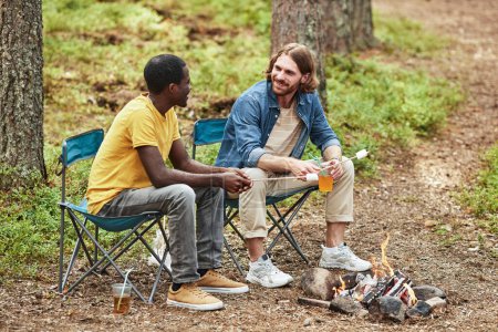 Téléchargez les photos : Des jeunes hommes assis sur des chaises, parlant entre eux et faisant frire des guimauves sur le feu de camp dans la forêt - en image libre de droit