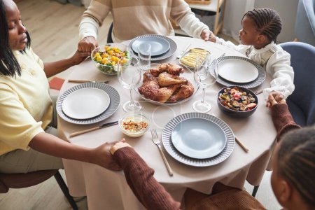 Téléchargez les photos : Vue en angle élevé de la famille africaine de quatre personnes assises à table les yeux fermés et priant en se tenant la main pendant le dîner - en image libre de droit