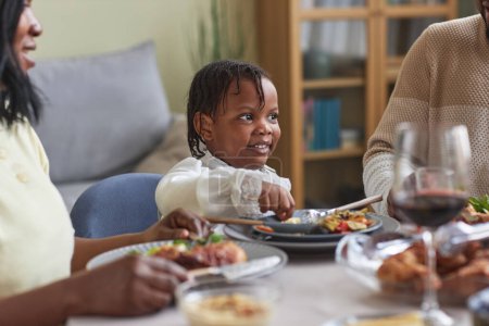 Téléchargez les photos : Petite fille africaine assise à table et dînant avec sa famille à la maison - en image libre de droit