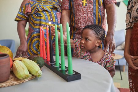 Foto de Niña africana encendiendo velas en la mesa mientras celebra las vacaciones de Kwanzaa junto con su familia - Imagen libre de derechos