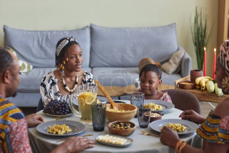 Téléchargez les photos : Famille africaine en costumes nationaux assis à table et parlant entre eux, ils célèbrent les vacances de Kwanzaa - en image libre de droit