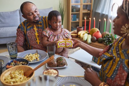 Photo for African parents giving present to their little daughter for holiday while they sitting at dining table - Royalty Free Image