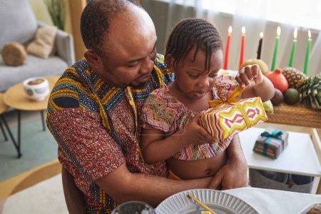 Téléchargez les photos : Petite fille africaine ouvrant boîte cadeau alors qu'elle était assise sur les genoux de son père à table - en image libre de droit