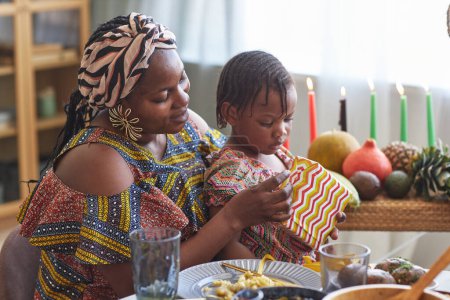 Téléchargez les photos : Femme africaine en costume national cadeau d'ouverture avec sa petite fille alors qu'ils sont assis à table - en image libre de droit