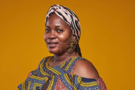 Foto de Retrato de mujer hermosa africana en elegantes pendientes y pañuelo para la cabeza sonriendo a la cámara sobre fondo amarillo - Imagen libre de derechos