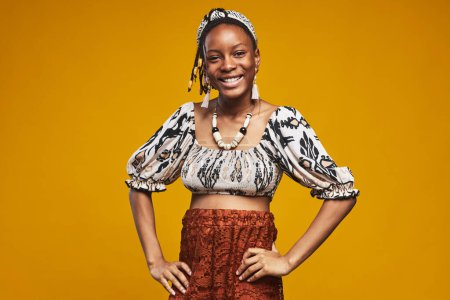 Téléchargez les photos : Portrait de fille africaine en vêtements nationaux souriant à la caméra debout sur fond jaune - en image libre de droit