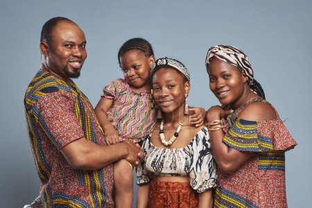 Téléchargez les photos : Portrait d'une heureuse famille africaine de quatre personnes vêtues de vêtements nationaux embrassant et souriant à la caméra debout sur fond bleu - en image libre de droit