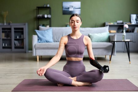 Téléchargez les photos : Jeune femme avec prothèse bras faire du yoga sur tapis d'exercice dans le salon - en image libre de droit