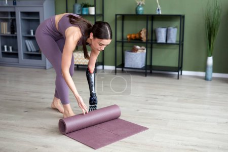 Téléchargez les photos : Jeune femme avec un bras prothétique finissant sa formation à la maison, tapis d'exercice de yoga roulant - en image libre de droit