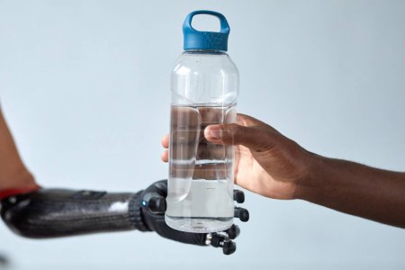 Téléchargez les photos : Gros plan de l'homme africain donnant une bouteille d'eau à une femme avec un bras prothétique isolé sur fond bleu - en image libre de droit