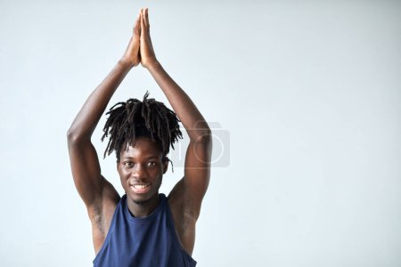 Téléchargez les photos : Portrait de jeune homme africain souriant à la caméra tout en faisant des exercices de yoga sur fond bleu - en image libre de droit