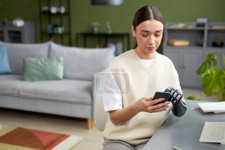 Foto de Chica joven con el brazo protésico usando su teléfono móvil mientras está sentada en la mesa en la sala de estar - Imagen libre de derechos