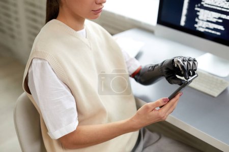 Téléchargez les photos : Gros plan de la jeune fille avec des codes de dactylographie de bras prothétiques sur smartphone tout en travaillant à son lieu de travail à la maison - en image libre de droit