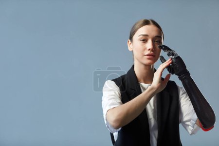Téléchargez les photos : Portrait de jeune femme avec bras prothétique regardant la caméra debout sur fond bleu - en image libre de droit