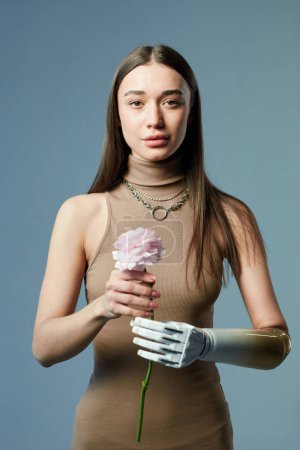 Téléchargez les photos : Portrait de jeune fille avec un bras prothétique tenant fleur et regardant la caméra sur fond bleu - en image libre de droit