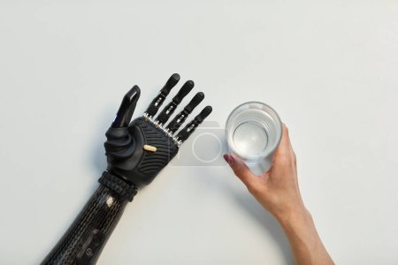 Photo for High angle view of young woman with prosthetic arm holding glass of water and drinking pill - Royalty Free Image