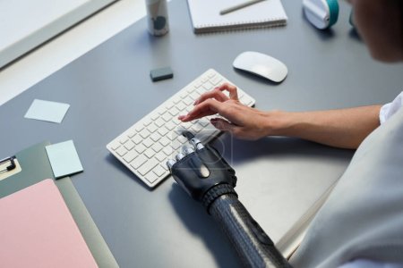 Téléchargez les photos : Gros plan de la femme avec un bras prothétique tapant sur le clavier pendant son travail sur ordinateur à la table - en image libre de droit
