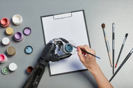 Téléchargez les photos : Vue grand angle de la femme avec peinture de bras prothétique avec peintures et pinceau sur papier à la table - en image libre de droit