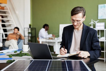 Téléchargez les photos : Jeune homme d'affaires prenant des notes dans un carnet tout en travaillant à table avec ordinateur portable et panneau solaire - en image libre de droit