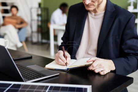 Téléchargez les photos : Gros plan d'un homme d'affaires écrivant des plans dans son carnet tout en travaillant à table sur un ordinateur portable - en image libre de droit
