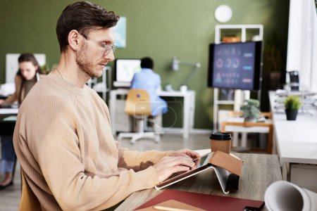 Téléchargez les photos : Homme d'affaires sérieux tapant sur tablette numérique et faisant son travail en ligne au travail au bureau - en image libre de droit