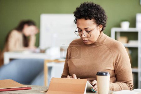 Foto de Empresaria africana concentrándose en su trabajo en línea en tableta digital mientras está sentada en la mesa de la oficina - Imagen libre de derechos