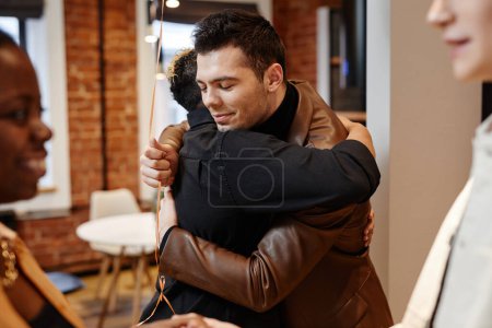 Foto de Feliz joven en chaqueta de cuero dando abrazo a la mujer negra en ropa de vaquero casualwear mientras la saluda en el pasillo del apartamento - Imagen libre de derechos