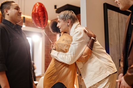 Téléchargez les photos : Vue latérale de deux jeunes amis en tenue décontractée s'embrassant tout en se tenant devant des personnes multiculturelles dans le couloir - en image libre de droit