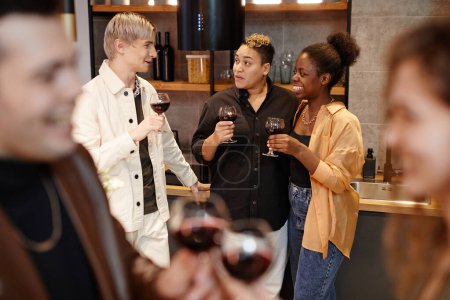 Téléchargez les photos : Groupe de jeunes amis interculturels avec des verres de vin rouge bavarder les uns avec les autres dans la cuisine tout en appréciant la fête à la maison - en image libre de droit
