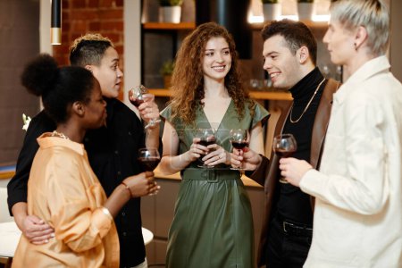 Téléchargez les photos : Groupe de jeunes amis multiculturels en tenue décontractée trinquant à la fête à la maison et bavardant debout l'un devant l'autre dans la cuisine - en image libre de droit