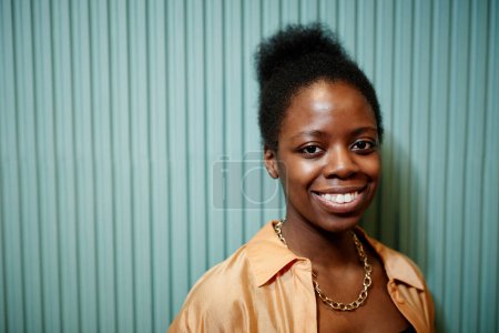 Téléchargez les photos : Jeune femme noire joyeuse en chemise décontractée jaune regardant la caméra avec le sourire de dents tout en se tenant debout sur fond bleu clair - en image libre de droit