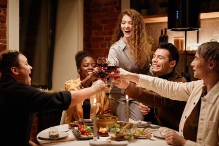 Téléchargez les photos : Jeunes amis extatiques clinquant avec du vin rouge dans des verres à vin sur la table du dîner servi avec de la nourriture maison et décoré de bougies allumées - en image libre de droit