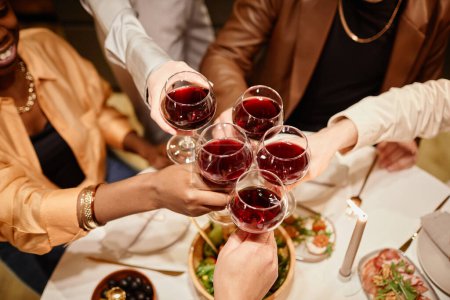 Foto de Vista superior de las manos de los jóvenes amigos tintineo con gafas de vino sobre la mesa de la cena servida con comida casera durante la celebración - Imagen libre de derechos