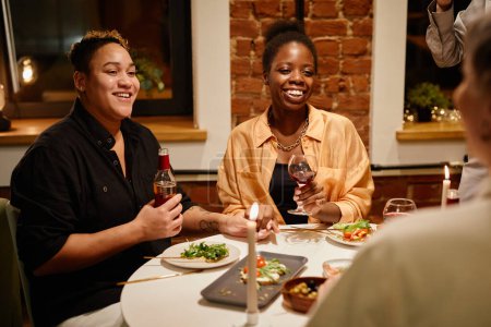 Foto de Feliz joven pareja lesbiana con bebidas alcohólicas sentada junto a la mesa servida y hablando con amigos que vinieron a cenar y a la fiesta en casa - Imagen libre de derechos
