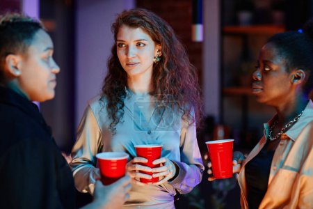 Foto de Grupo de mujeres jóvenes interculturales con bebidas en copas rojas charlando mientras disfrutan de la reunión o la fiesta en casa en la noche de fin de semana - Imagen libre de derechos