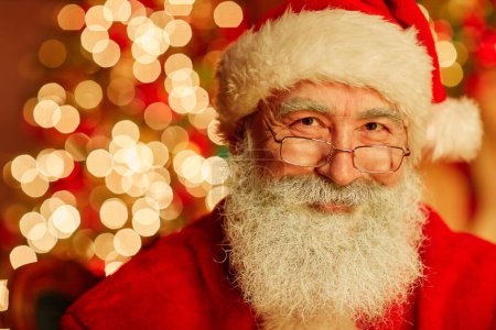 Téléchargez les photos : Portrait rapproché du Père Noël traditionnel regardant la caméra et souriant avec des lumières de Noël scintillantes en arrière-plan, espace de copie - en image libre de droit